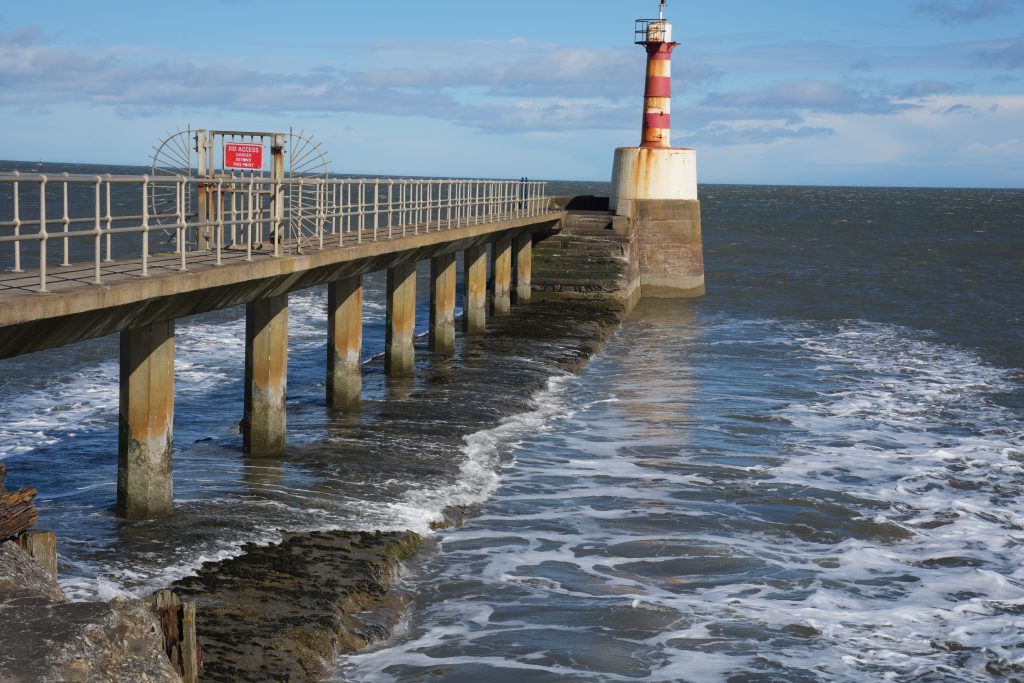 Pier and lighthouse at Amble Harbour in Northumberland, with waves rolling against the sea wall beneath blue skies along the northeast coastline.
