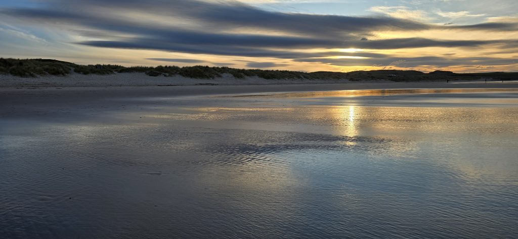 Sunset over Bamburgh Beach in Northumberland, with soft golden light reflecting across wet sand, rolling dunes and calm coastal water beneath dramatic evening clouds.