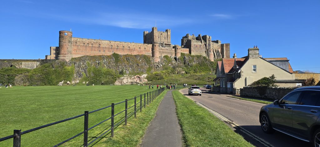 Bamburgh Castle rising above the Northumberland coastline on a bright spring day, viewed from the village road with green fields, historic stone walls and blue skies surrounding the fortress.