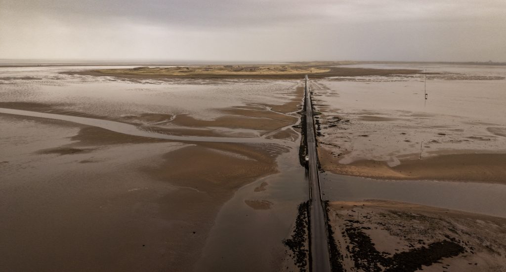 Aerial view of the tidal causeway leading to Holy Island in Northumberland, surrounded by sweeping sand flats, shallow tidal waters and muted coastal tones beneath overcast skies.