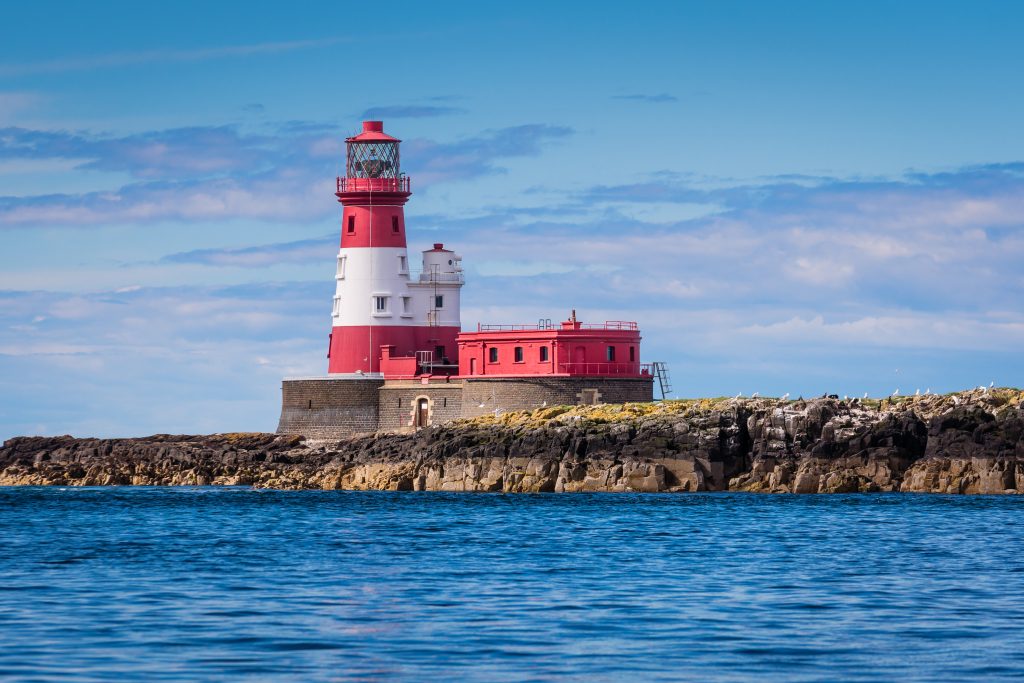 Longstone Lighthouse near the Farne Islands off the Northumberland coast, standing on rocky shoreline beneath bright blue skies and surrounded by calm sea water.