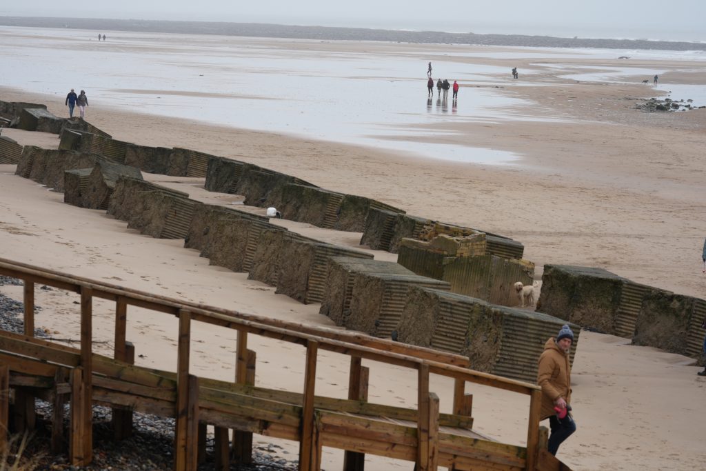 People walking across a wide sandy beach in Northumberland beside weathered sea defences and a wooden boardwalk on an overcast coastal day.