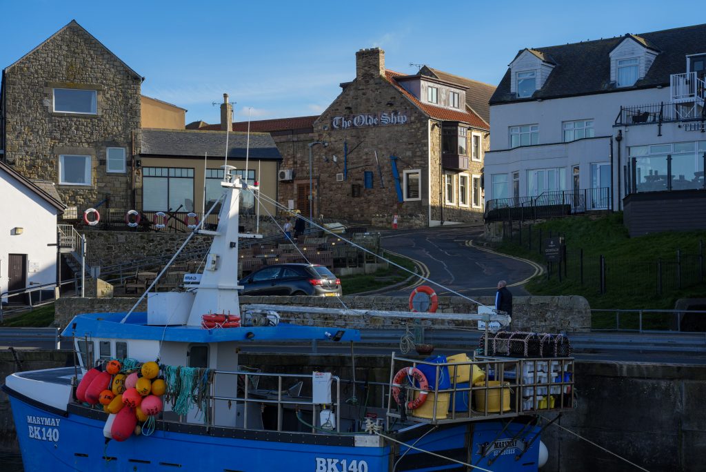 Fishing boat moored in Seahouses harbour in Northumberland beside traditional stone buildings and The Olde Ship Inn beneath clear blue evening skies.