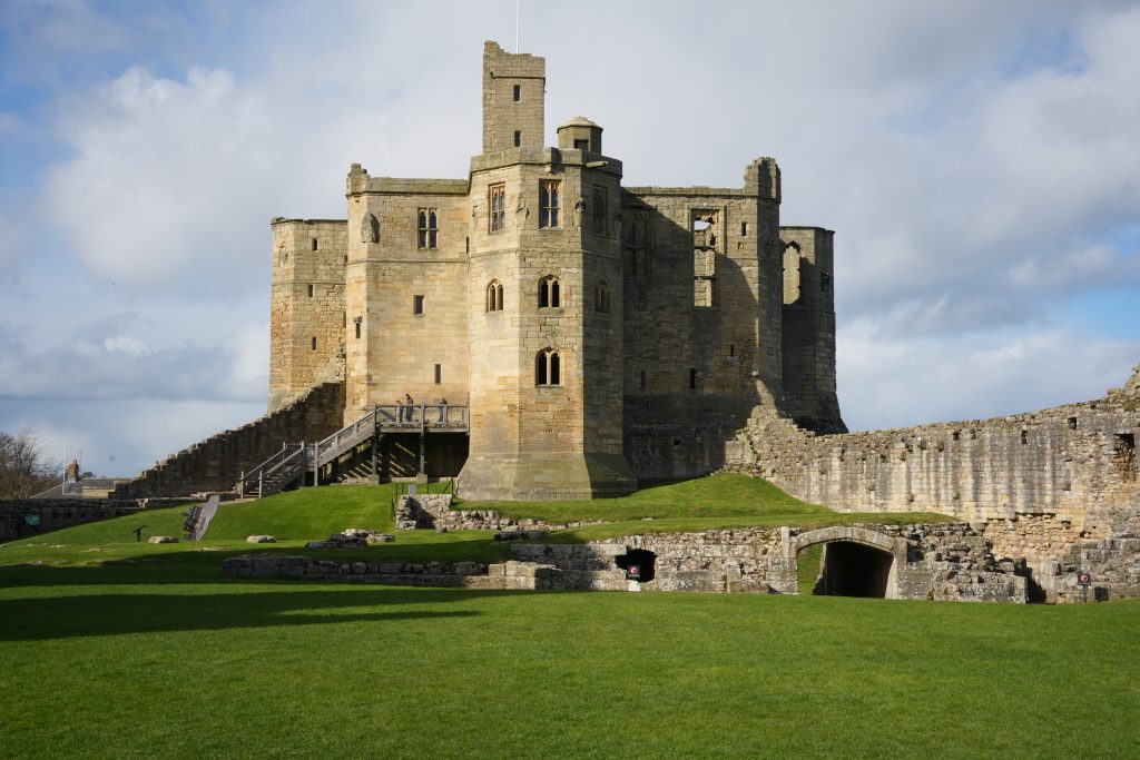 Warkworth Castle in Northumberland standing above green grounds beneath dramatic clouds, showcasing medieval stone towers, fortified walls and historic English architecture.